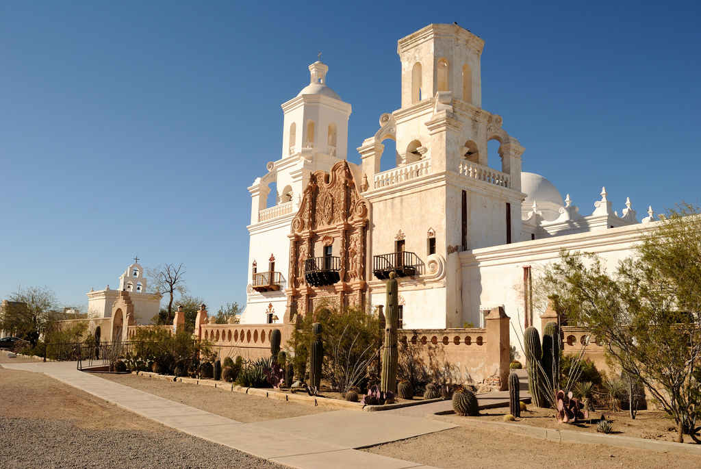 View along the cactus-studded front of the mission with a smaller outbuilding with three bells visible.