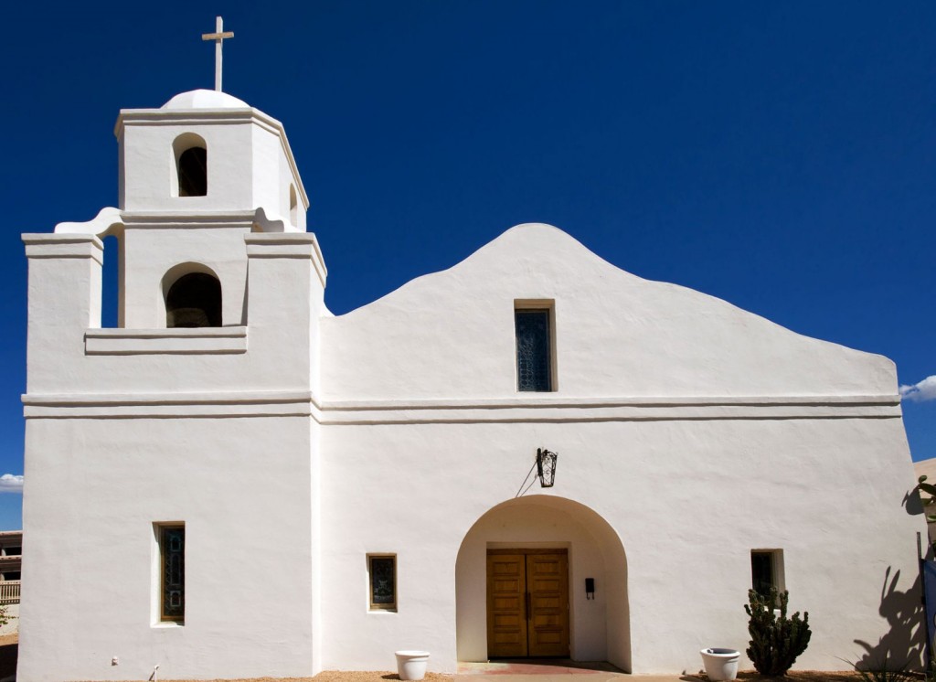 Front of a historic white adobe mission with narrow windows, a wooden door, and a belltower.