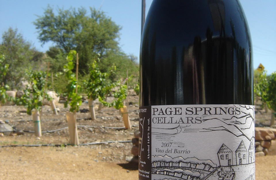 A dark bottle with a label reading Page Springs Cellars in the foreground while vines are visible in the background.