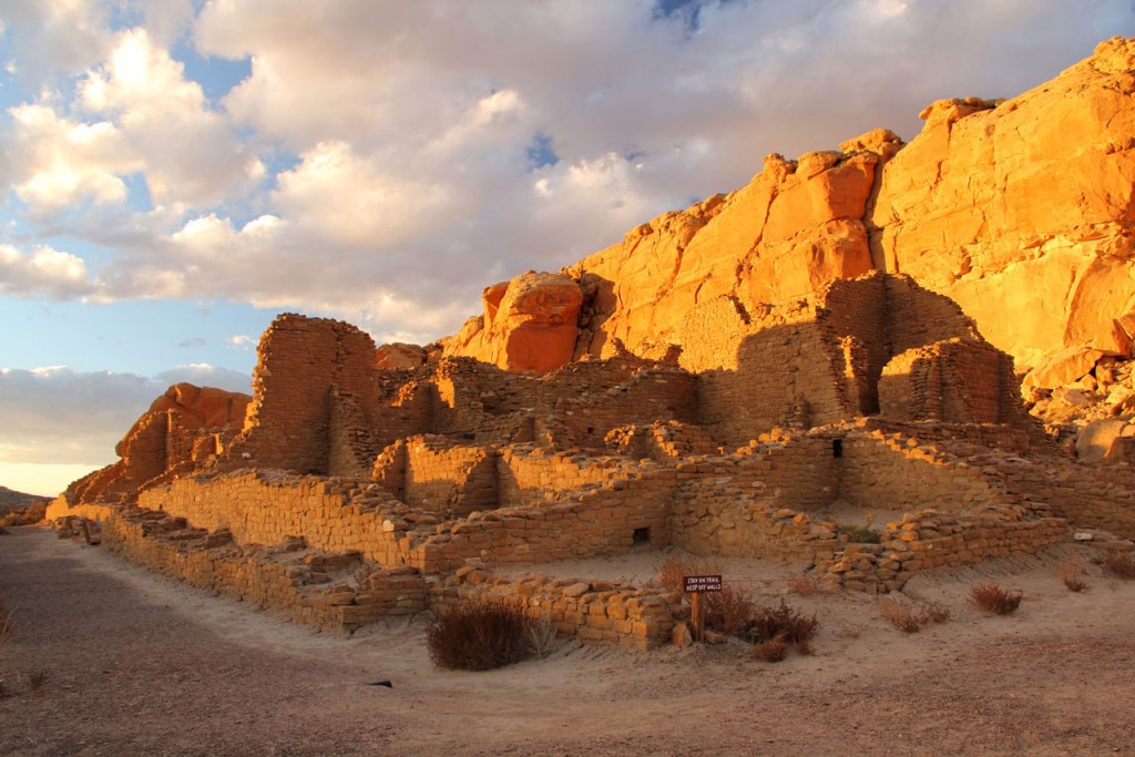 Afternoon sun turns the rocks orange behind the remains of Pueblo ruins.