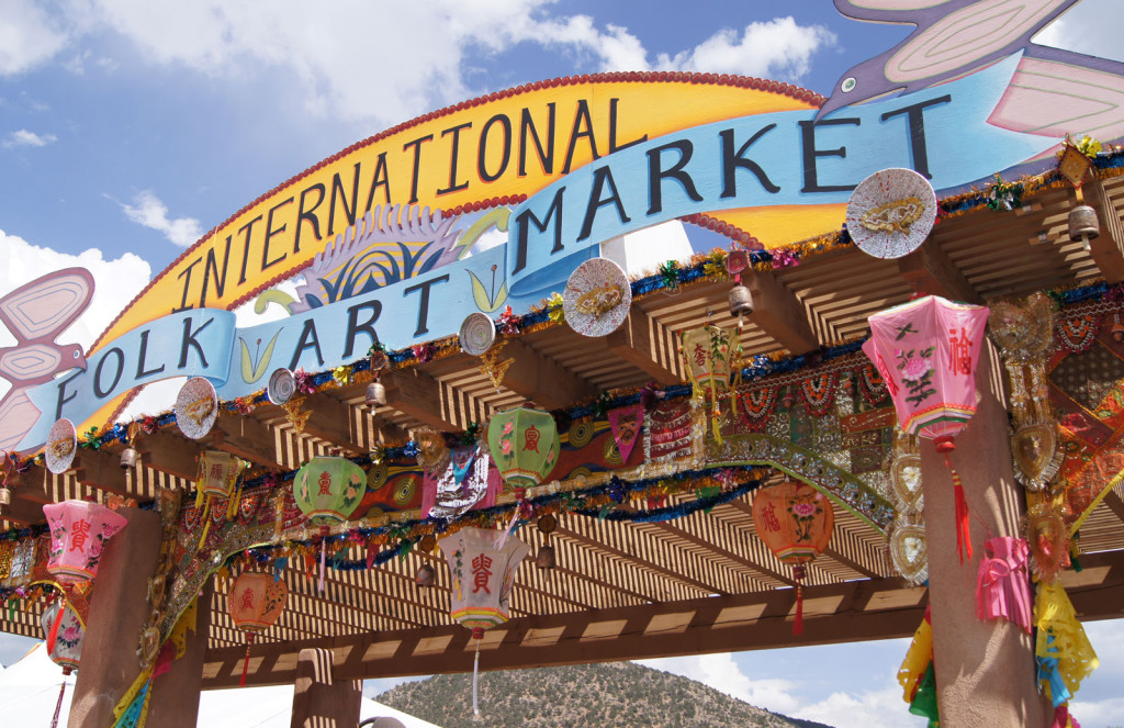 Open wooden entrance hung with colorful paper lanterns and a painted sign reading International Folk Art Market.