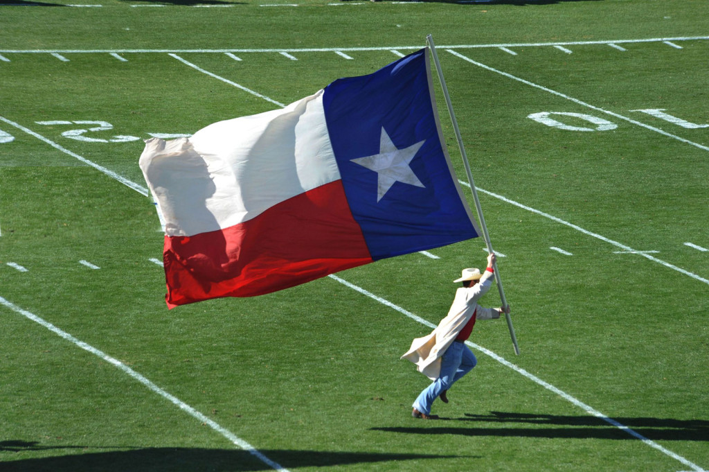 A man runs across a football field carrying the state flag which is nearly as tall as himself.