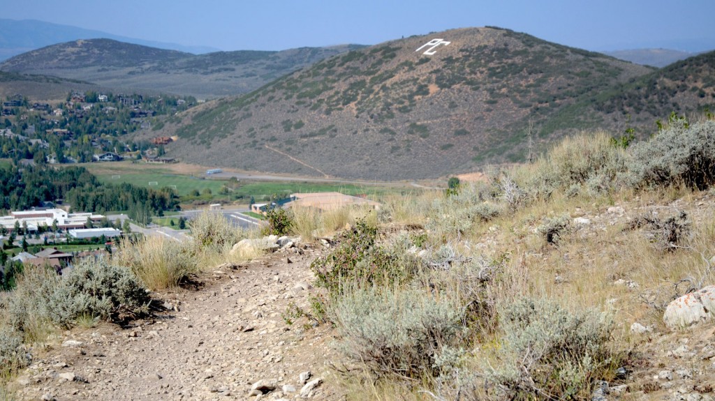 Hilltop view of Park City and surrounding hills, one with PC spelled out on the hillside. Short wild grasses grow in the foreground.