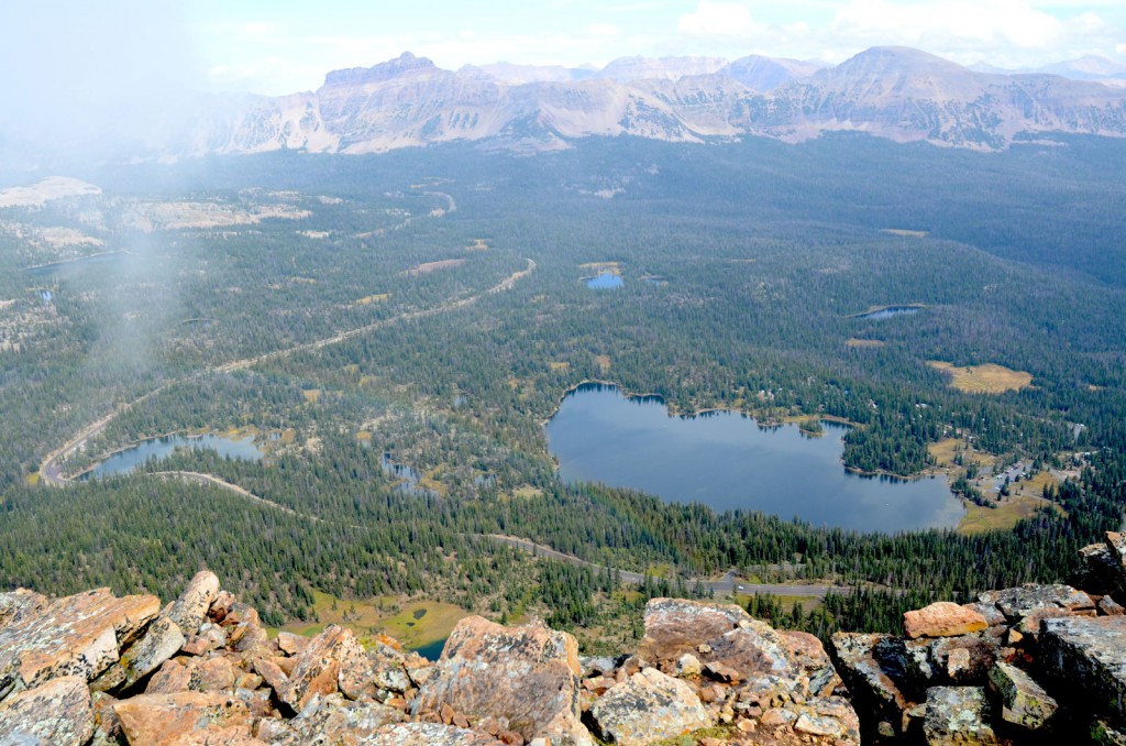 Large fragments of rock form a ledge in the foreground while a wide forested vista includes a large lake.