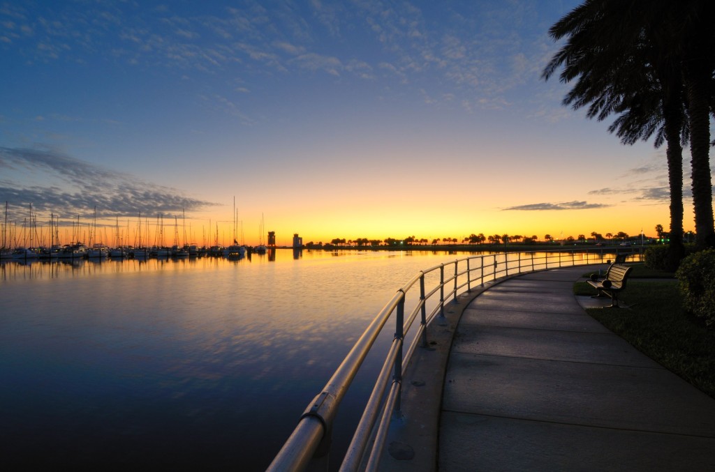 A walkway curves along the waterfront with the rising sun reflecting in the calm water and silhouetted palm trees lined up in the distance.