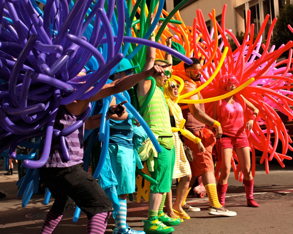 Six people standing in a row each wearing a different color of the rainbow, their costumes made out of long noodle balloons.