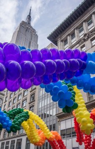 Looking up at rainbow balloons strung in arches on a street with the Empire State Building in the background. 