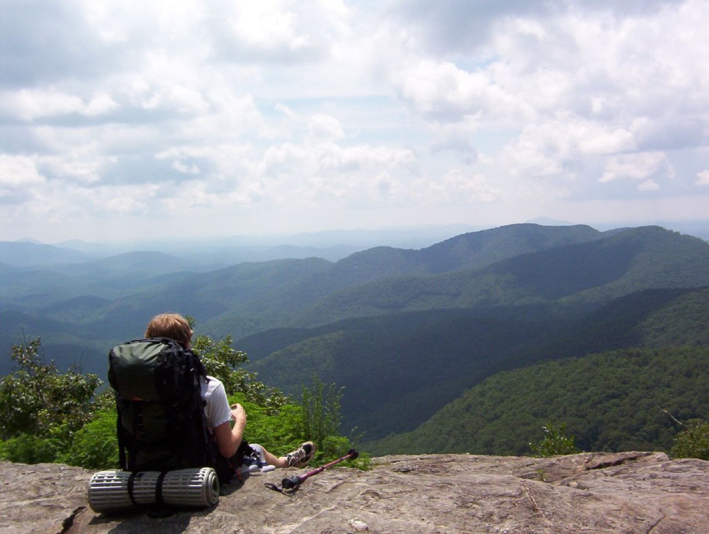 A hiker with a backpack sits on a bare patch of stone staring out over the mountainous landscape.