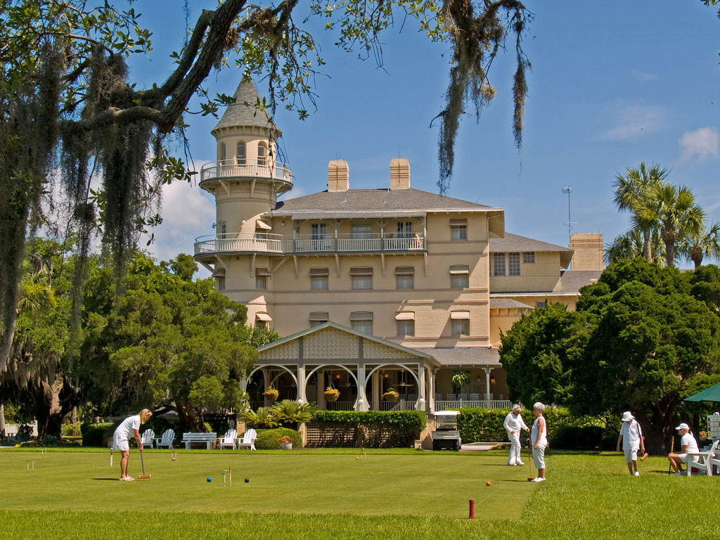 Patrons dressed all in white play croquet on the lawn in front of the multistory colonial club building.