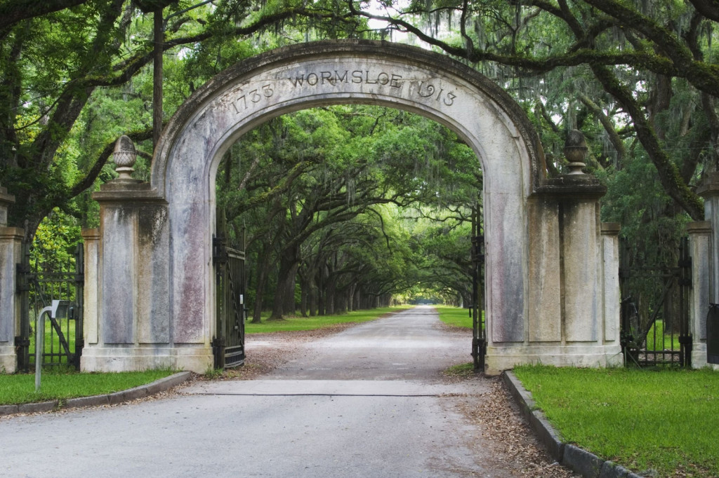 A stone arch with Wormsloe carved into it rises over a tree-lined road.