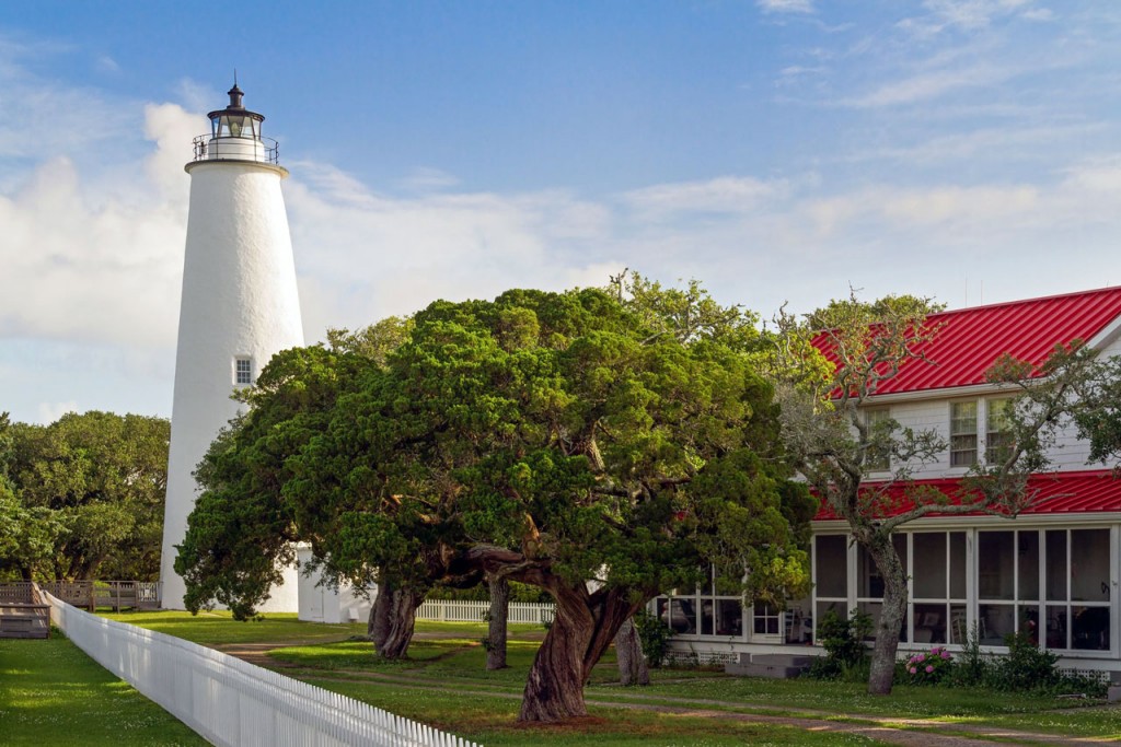 View of a squat white-washed lighthouse with a two-story keeper's house surrounded by a white picket fence.