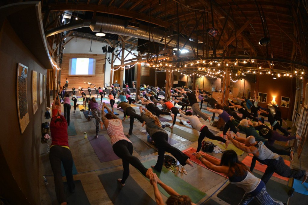 A large room is filled with about a hundred people practicing yoga together.