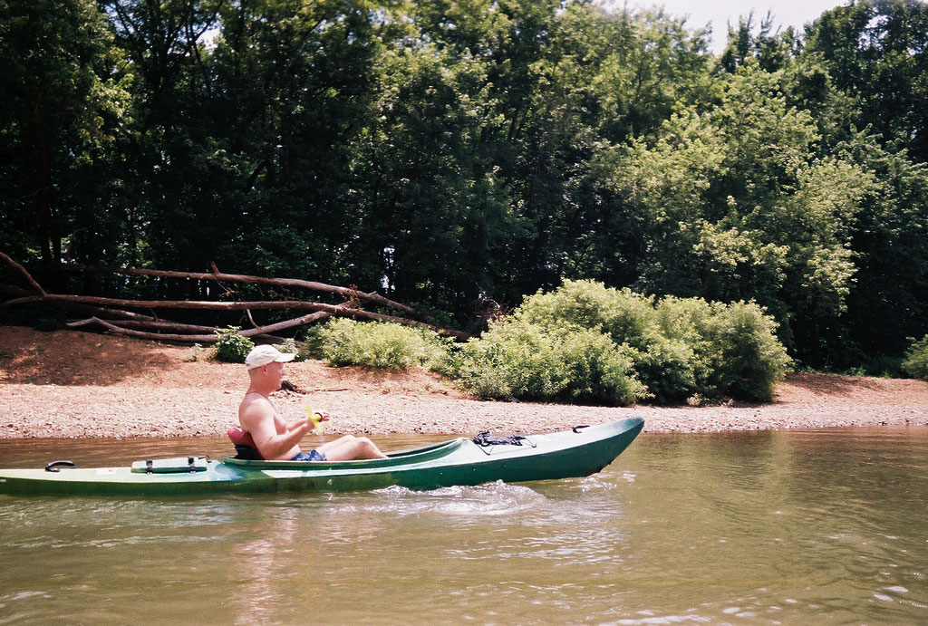 A man paddles a kayak in shallow water near a rocky shore.