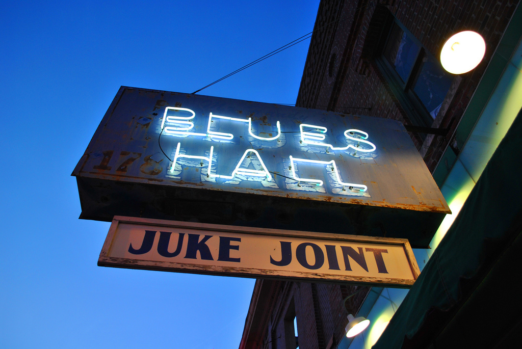 View up at a sign at dusk with Blues Hall written in blue neon and a smaller Juke Joint sign hanging below it.