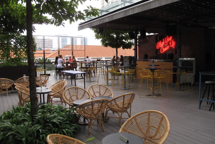 Wicker chairs clustered around tables at a terrace bar, with trees and greenery in the background