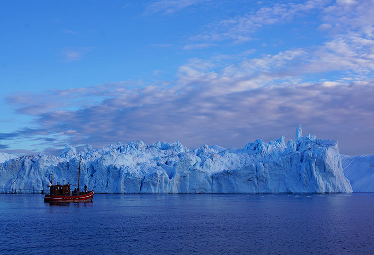 A red sailing boat is dwarfed by a craggy chunk of the Ilulissat glacier, a blue outcrop in the waters of Disko Bay, Greenland