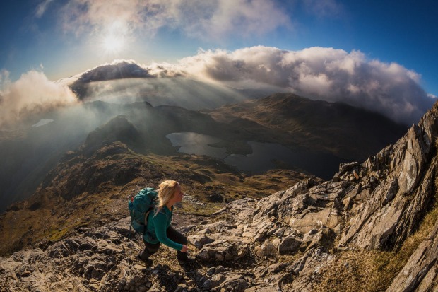 A young woman walking on the rocky slopes of Crib Goch, and the view of the Llyn Llydaw lake and valley through low cloud.