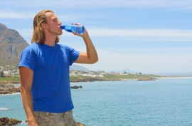 man in a blue t-shirt drinking water standing next to the ocean